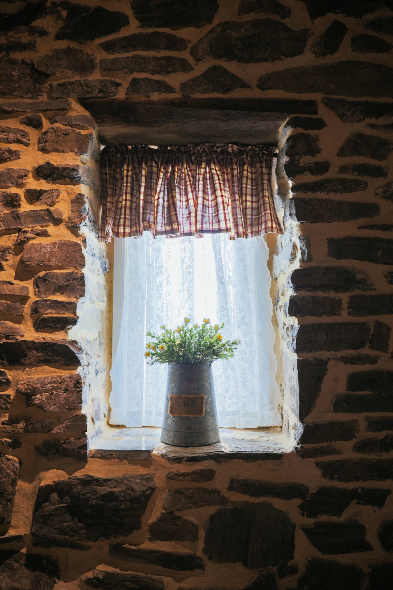 A deep-set window punched through a fieldstone wall, a striped valance, a sheer curtain, and a galvanised bucket of yellow flowers on the sill