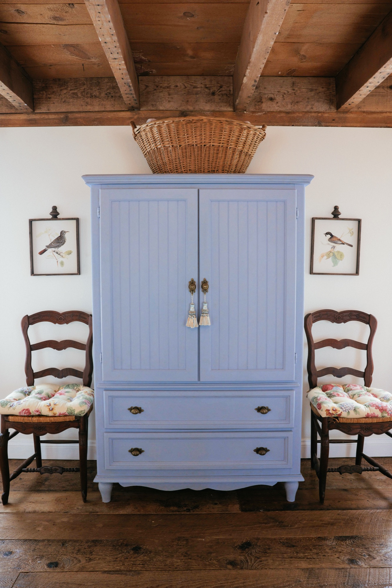 A periwinkle painted armoire flanked by two French ladderback chairs with chintz cushions and bird prints.