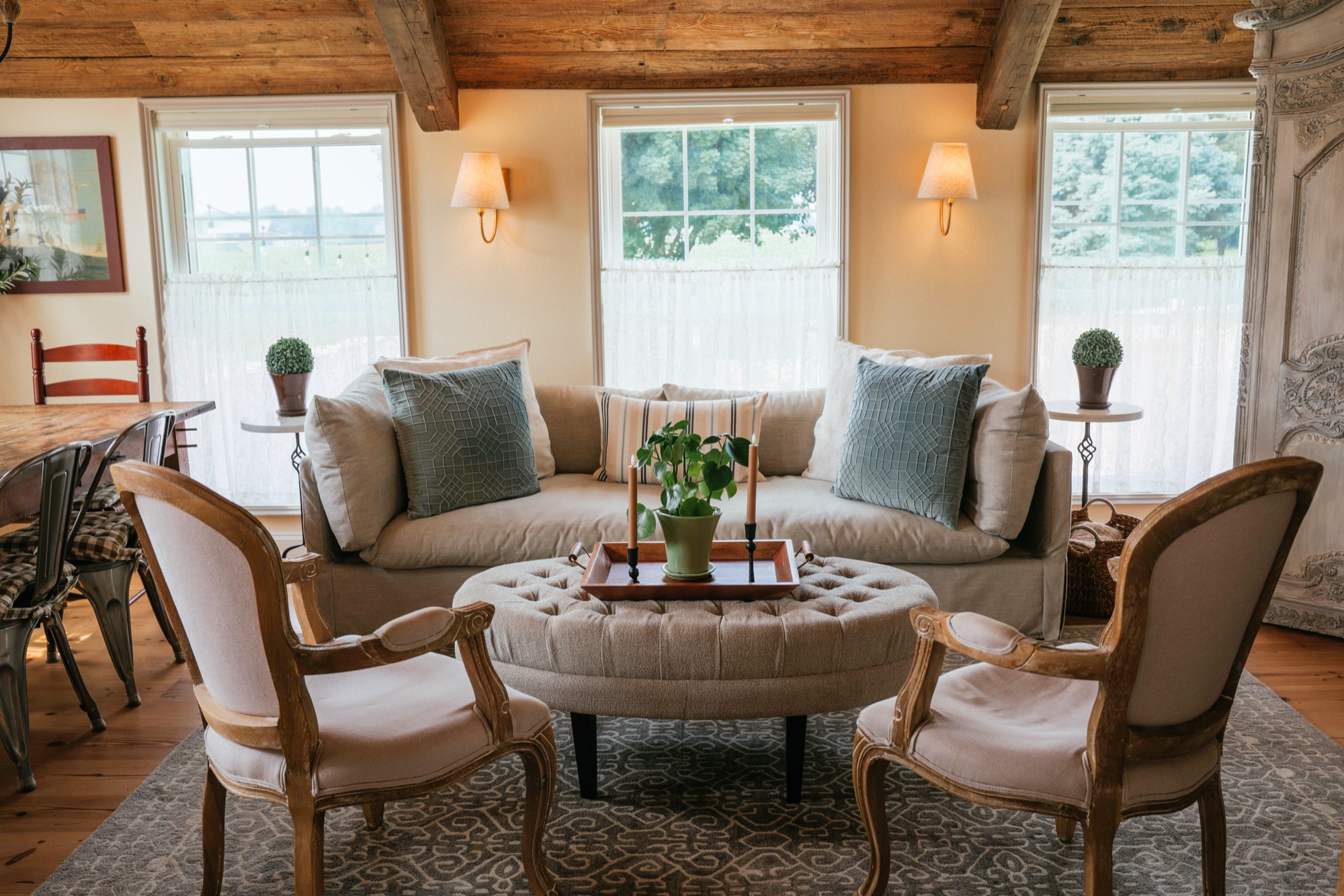 Living room with a linen sofa and carved armchairs under a hewn beam ceiling, three windows onto green foliage