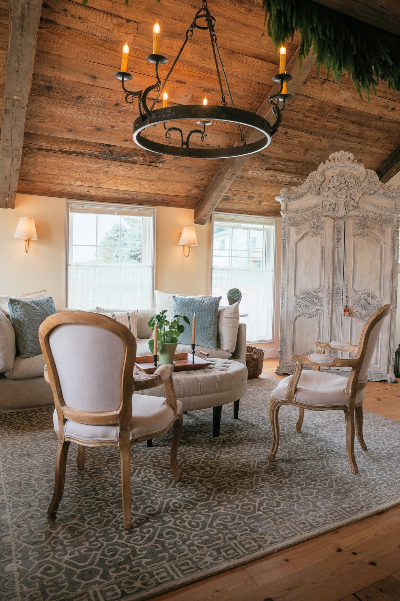 The salon: a linen sofa, two carved French armchairs and a tufted ottoman under a wrought-iron chandelier and hewn beams.