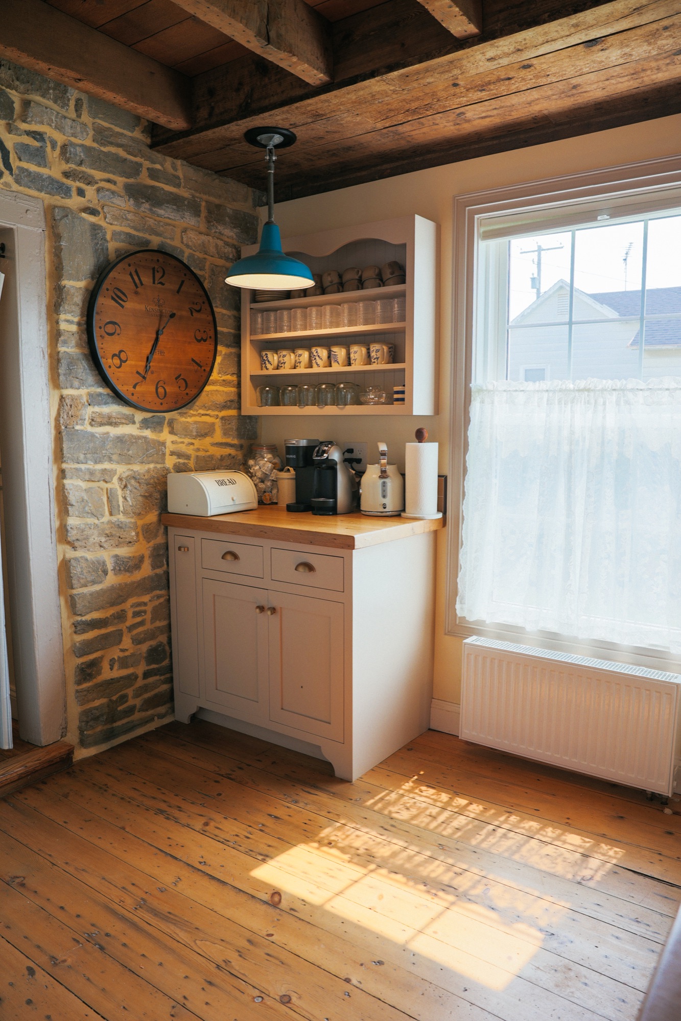 Coffee corner against a fieldstone wall, a round wooden clock, and wide pine floorboards in raked window light