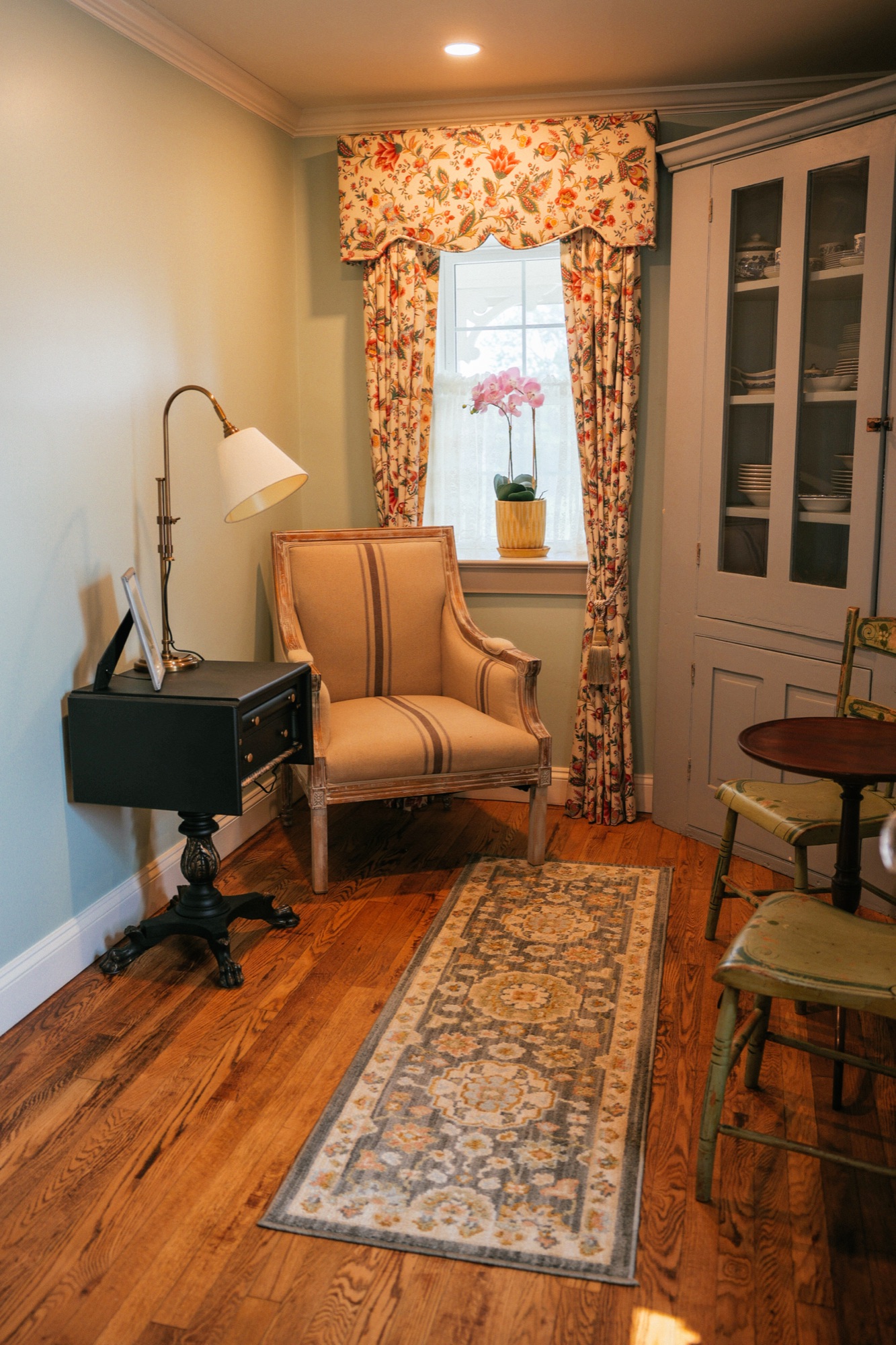 A quiet corner: a tan linen French armchair by a window with a floral valance, a brass reading lamp and a runner.