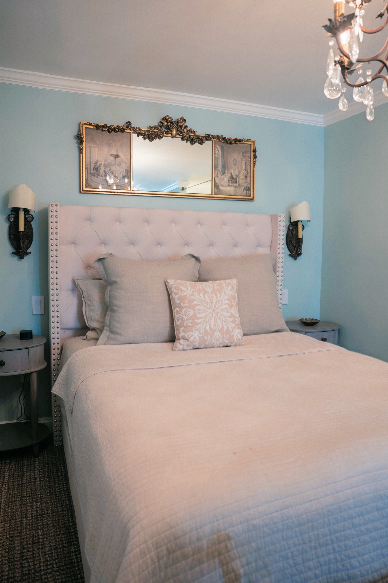 A pale blue bedroom with a gilt trumeau mirror over a tufted linen headboard and a crystal chandelier.