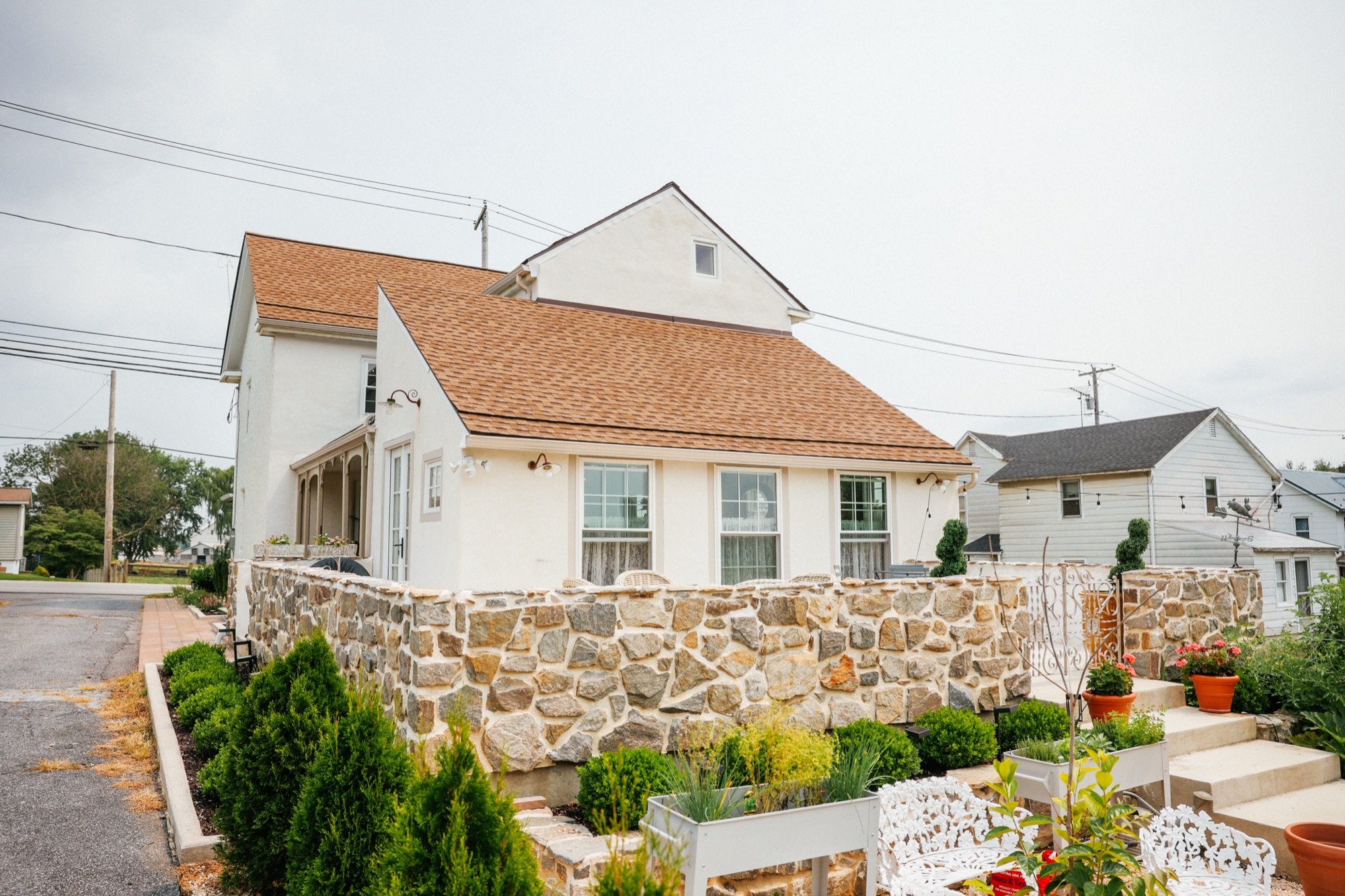 A long fieldstone retaining wall running the width of the frame, with the white stucco cottage and its brown shingle roof above it