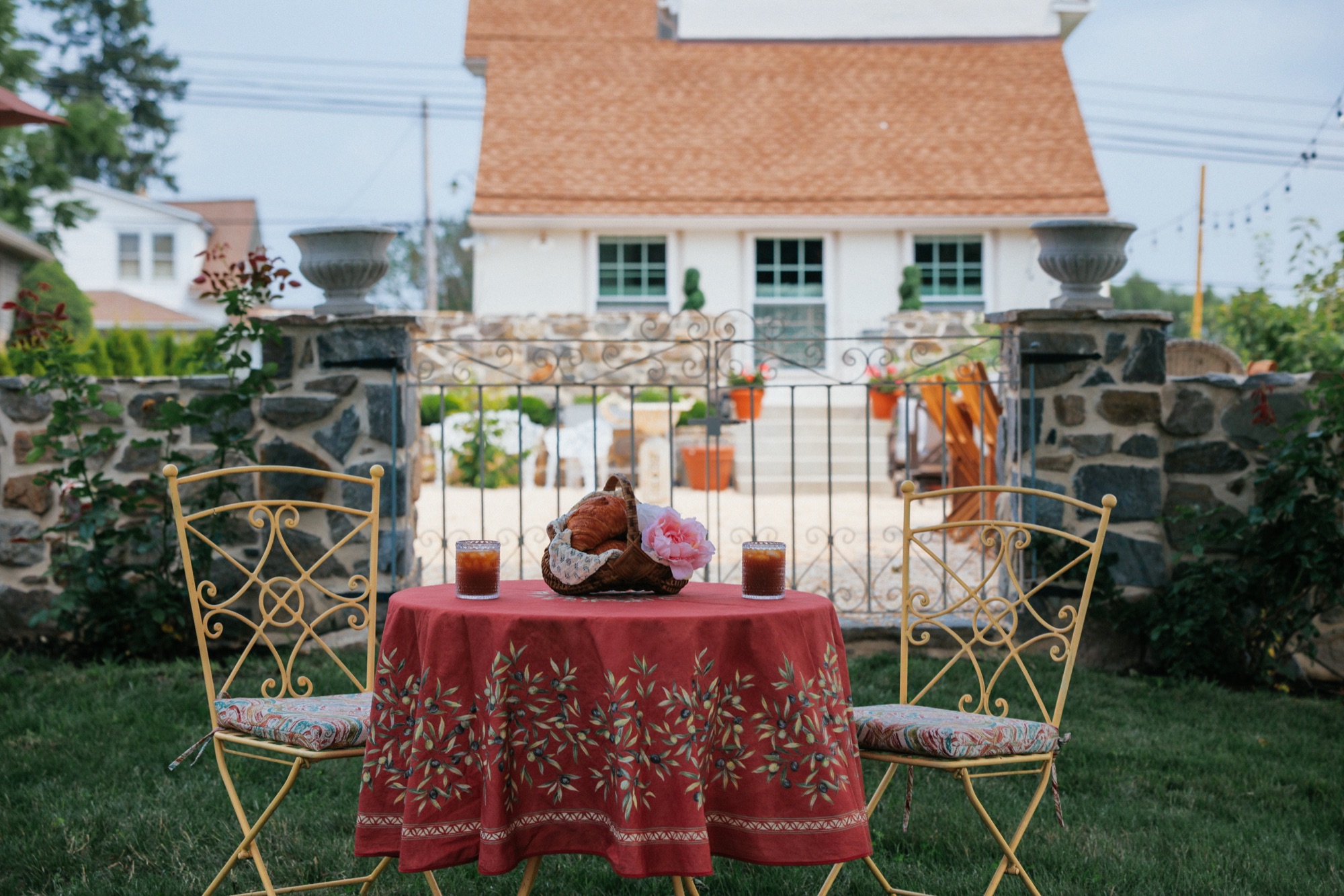 A round table under a madder-red Provencal olive-print cloth with two yellow wrought-iron bistro chairs, croissants and candles, the cottage behind.